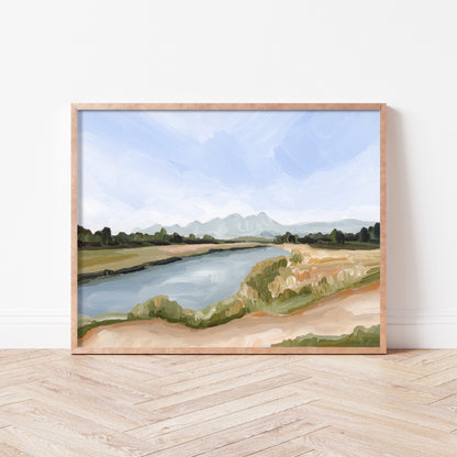 Framed landscape painting depicting a river winding through grassy terrain with mountains in the distance.