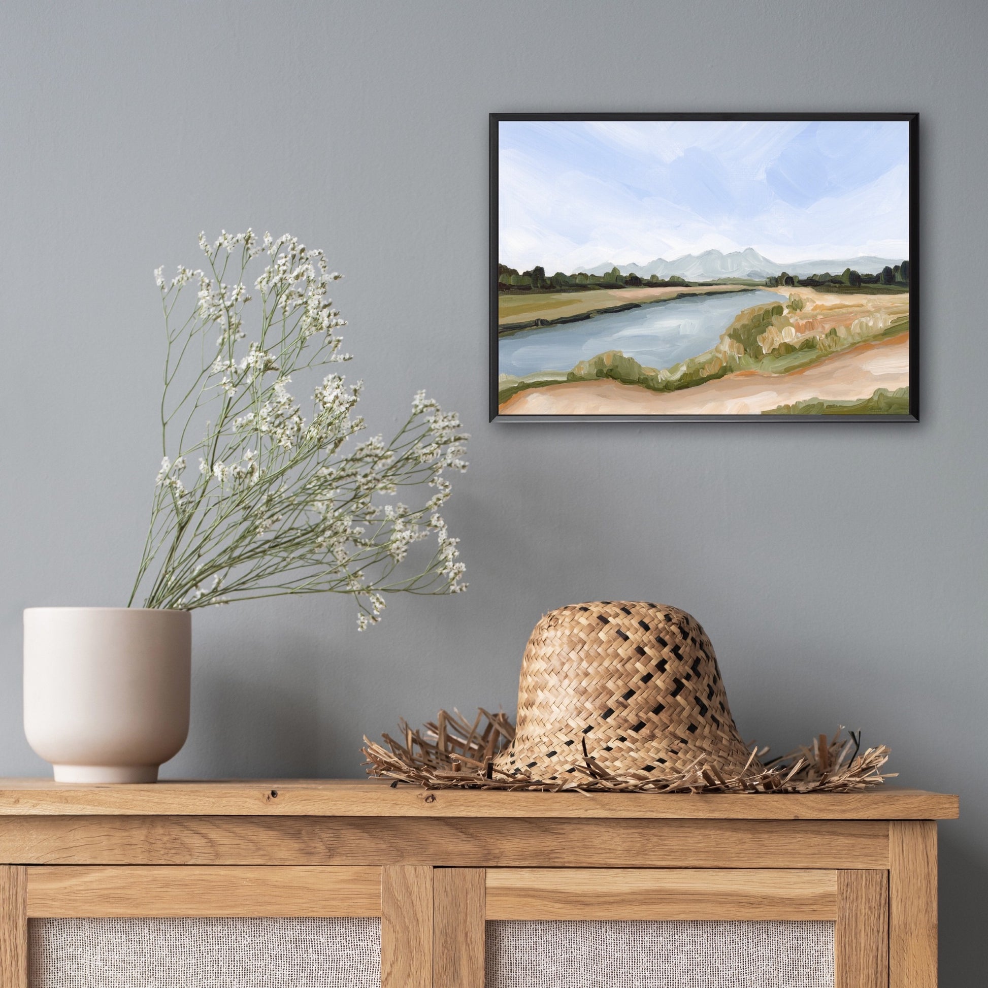 Framed landscape painting of a river and countryside hanging above a wooden console table with decorative items.