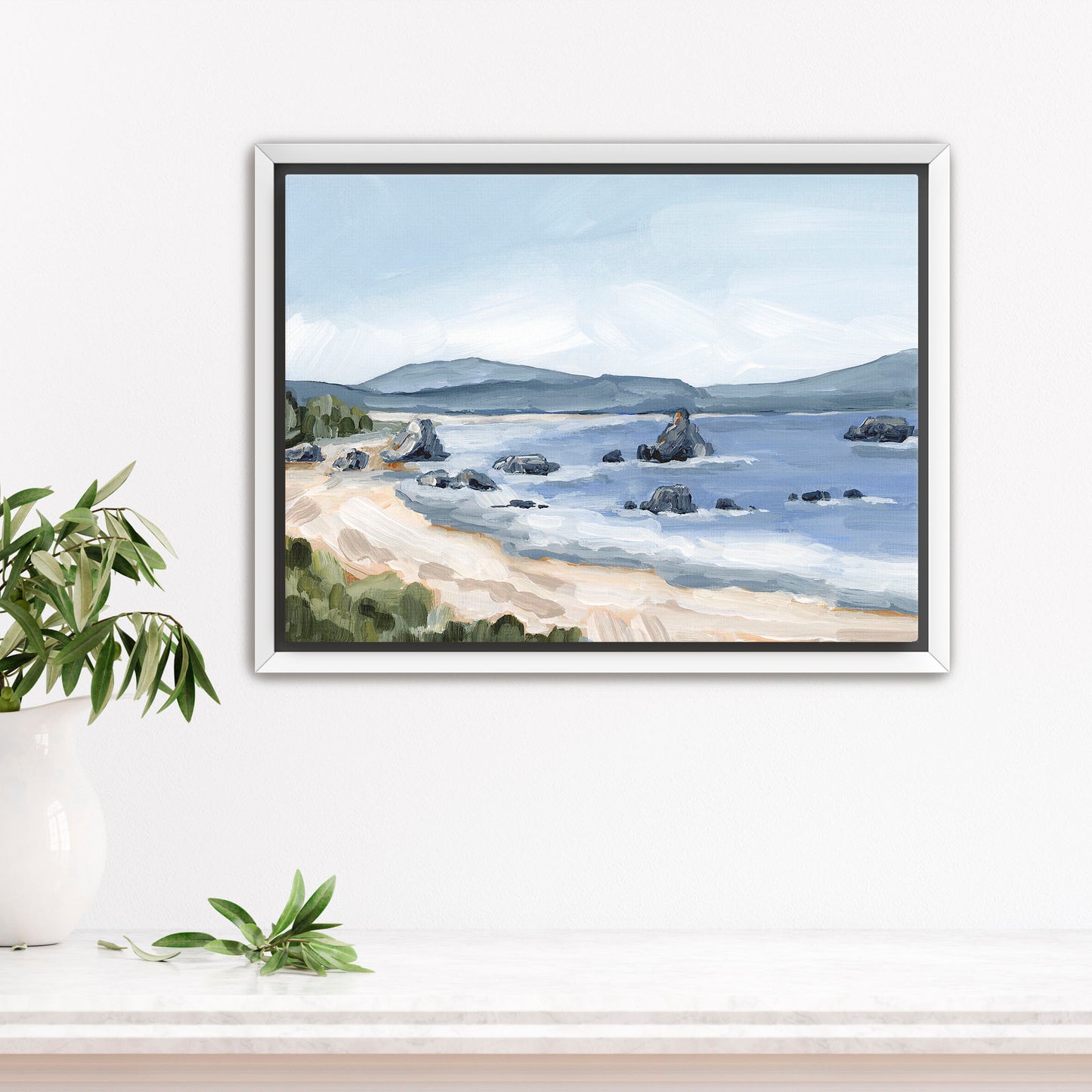 Framed painting of a coastal landscape with a beach, rocky shoreline, and distant mountains.