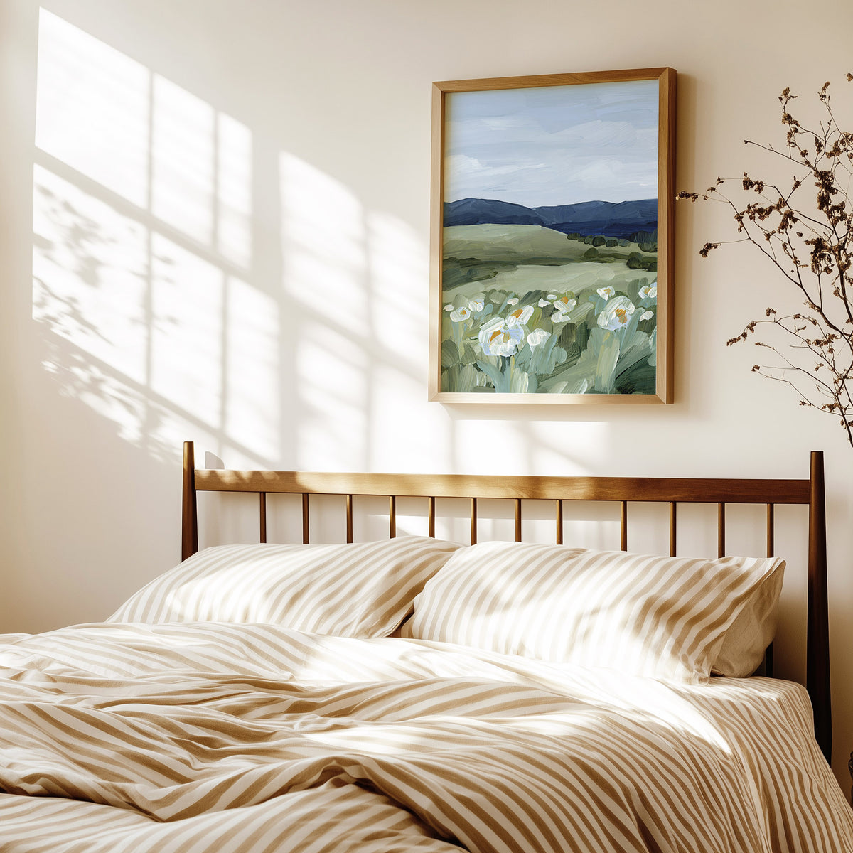 A framed landscape painting of white flowers in a green field hangs on a warm beige wall above a bed with beige and white striped bedding, styled with soft window shadow light.