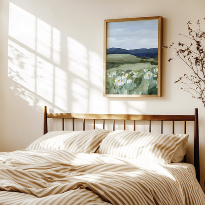 A framed landscape painting of white flowers in a green field hangs on a warm beige wall above a bed with beige and white striped bedding, styled with soft window shadow light.