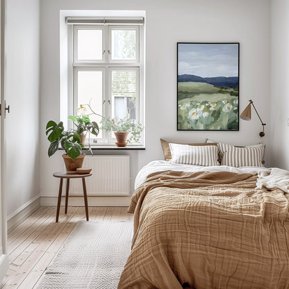 A styled bedroom interior with a bed dressed in beige bedding, striped pillows, a window with plants on the sill, and a framed landscape painting on the wall.