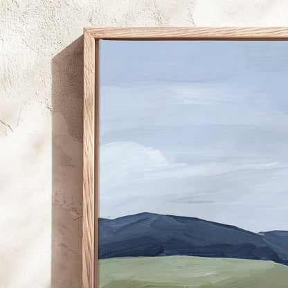 A framed landscape painting featuring a pale blue sky, dark blue mountains, and a light green field, set in a light wood frame against a textured beige plaster wall.