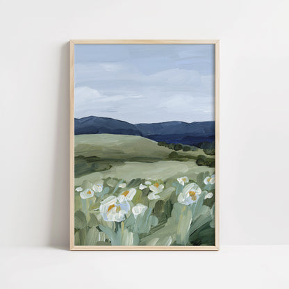 A framed landscape art print featuring white wildflowers in the foreground, green rolling hills, and dark blue mountains under a cloudy sky, displayed against a clean white wall.