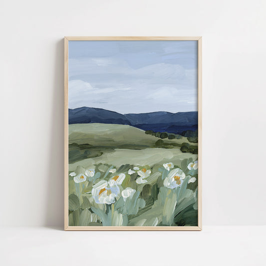 A framed landscape art print featuring white wildflowers in the foreground, green rolling hills, and dark blue mountains under a cloudy sky, displayed against a clean white wall.