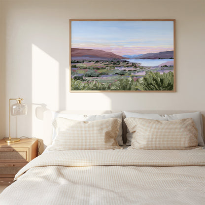 A framed landscape painting of a coastal scene with green shrubs, distant hills, and a pale sky, displayed above a neatly made bed in a softly lit bedroom.