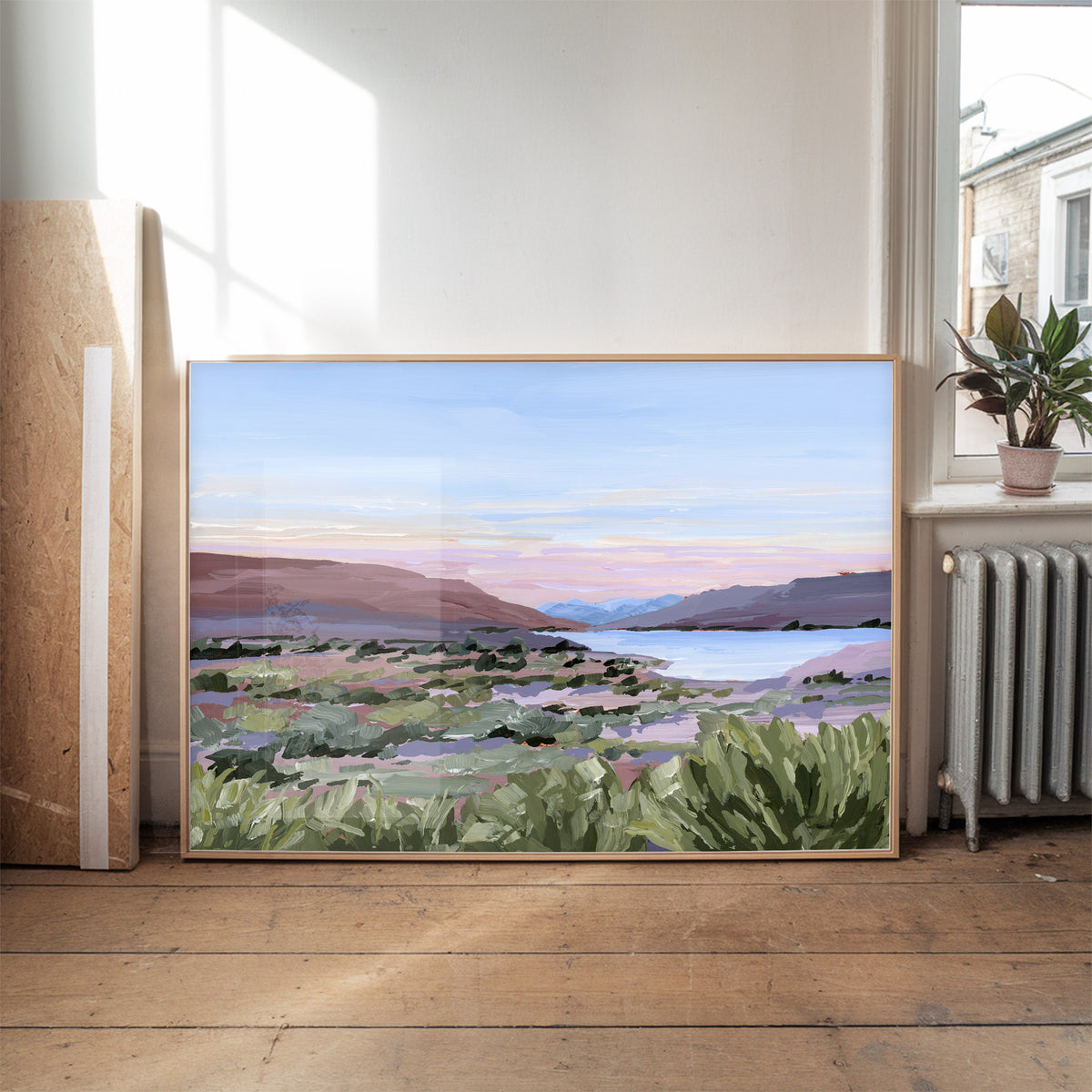 A framed landscape painting of a scenic valley with mountains, a lake, and green foreground foliage, leaning against a wall in a sunlit room with a wooden floor and a potted plant nearby.