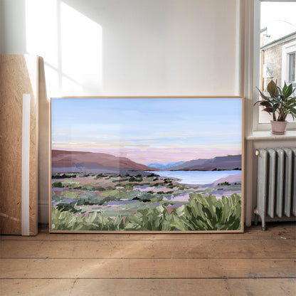 A framed landscape painting of a scenic valley with mountains, a lake, and green foreground foliage, leaning against a wall in a sunlit room with a wooden floor and a potted plant nearby.