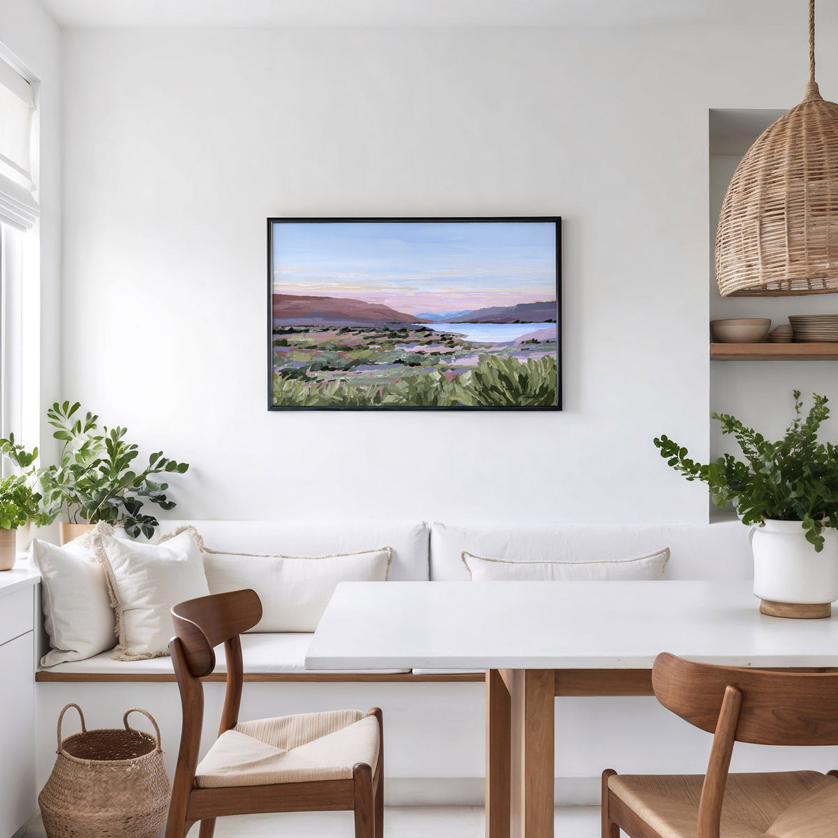 A framed landscape art print hangs on a white wall above a white bench and dining table in a bright, modern room with wooden chairs and potted plants.