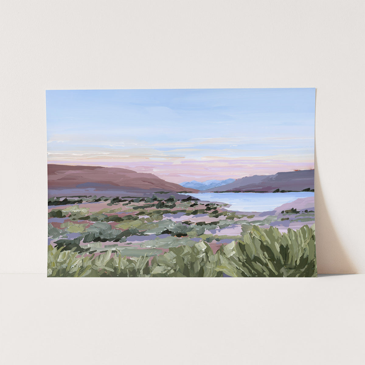 A landscape art print depicting a coastal scene with green shrubs in the foreground, distant mountains, and a pastel sky, shown leaning against a light beige wall.