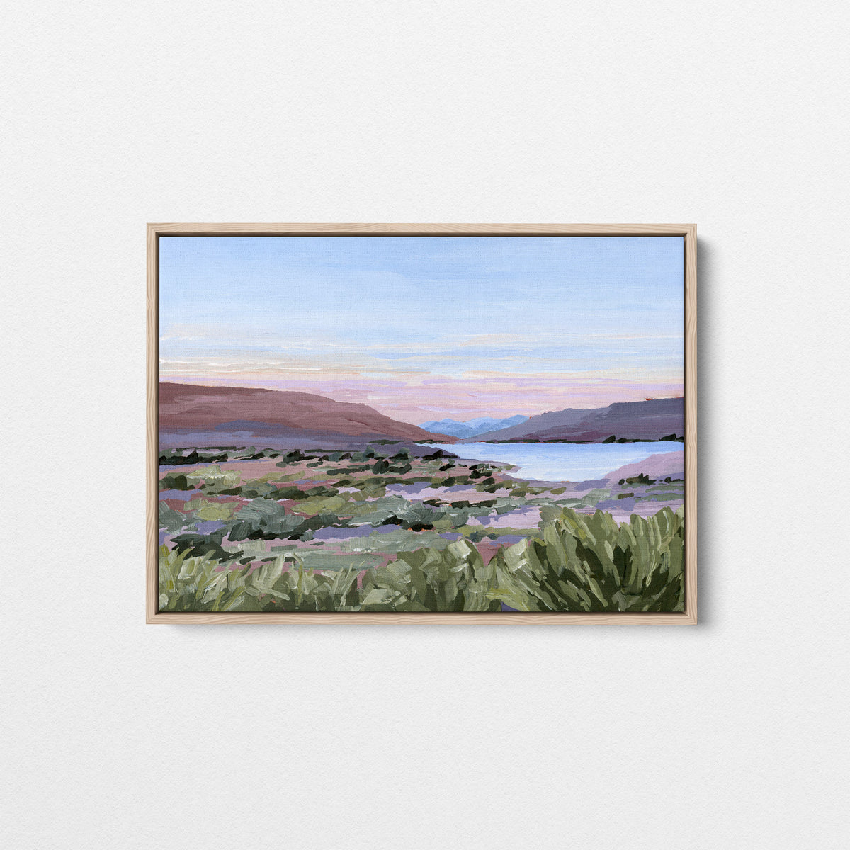 A framed landscape painting featuring green shrubs in the foreground, distant mountains, and a soft pink and blue sky, displayed on a clean white wall.