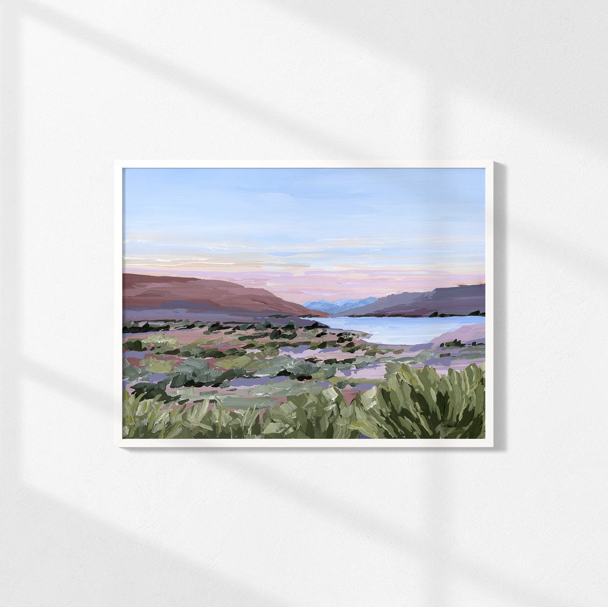 A framed landscape art print featuring a painted desert valley with green shrubs, distant mountains, and a soft blue and pink sky, displayed on a white wall with soft diagonal shadows.