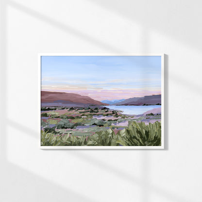 A framed landscape art print featuring a painted desert valley with green shrubs, distant mountains, and a soft blue and pink sky, displayed on a white wall with soft diagonal shadows.