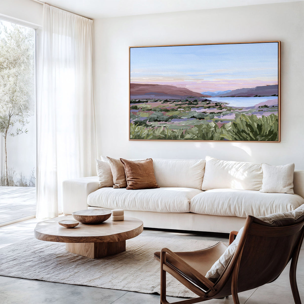 A large framed landscape painting of a coastal valley at sunset hangs on a white wall above a cream sofa in a bright, modern living room.