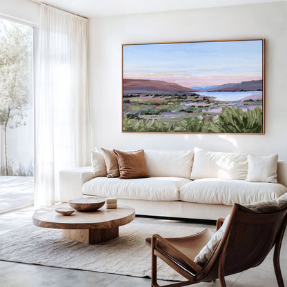 A large framed landscape painting of a coastal valley at sunset hangs on a white wall above a cream sofa in a bright, modern living room.