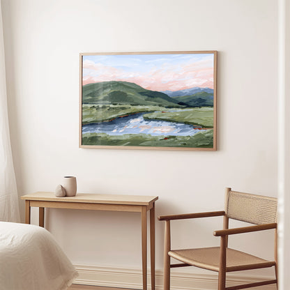A framed landscape painting of green hills, a lake, and a pink sunset sky, displayed on a white wall above a light wood console table and a woven-back chair.