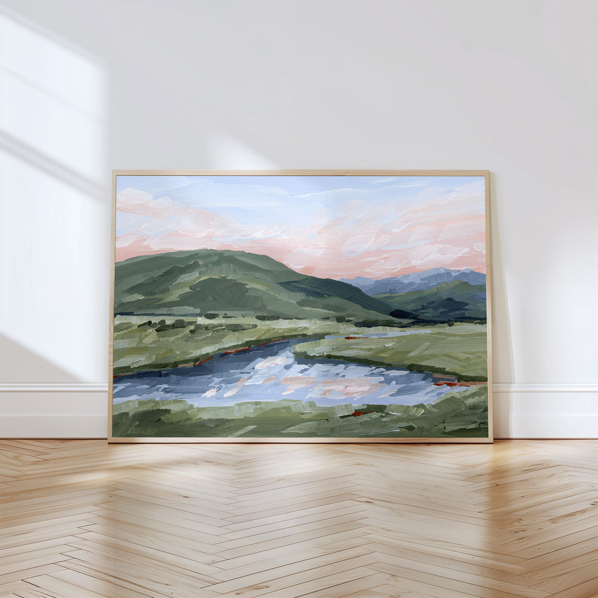 A framed landscape painting of green hills, a winding river, and a pink sky, displayed on a light wood floor against a white wall with soft window light.