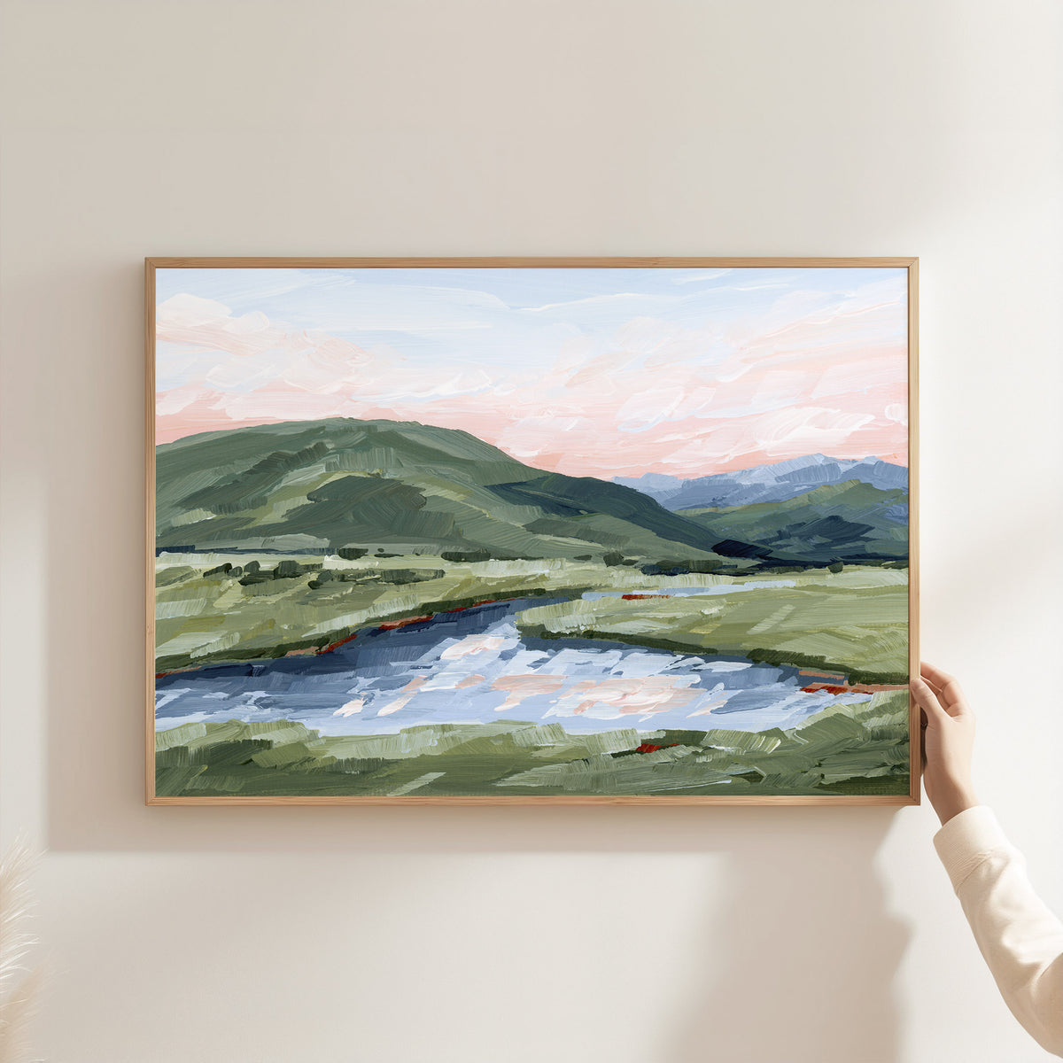 A framed landscape painting of green hills and a winding river under a soft pink and blue sky, mounted on a light beige wall with a hand holding the right side of the frame.