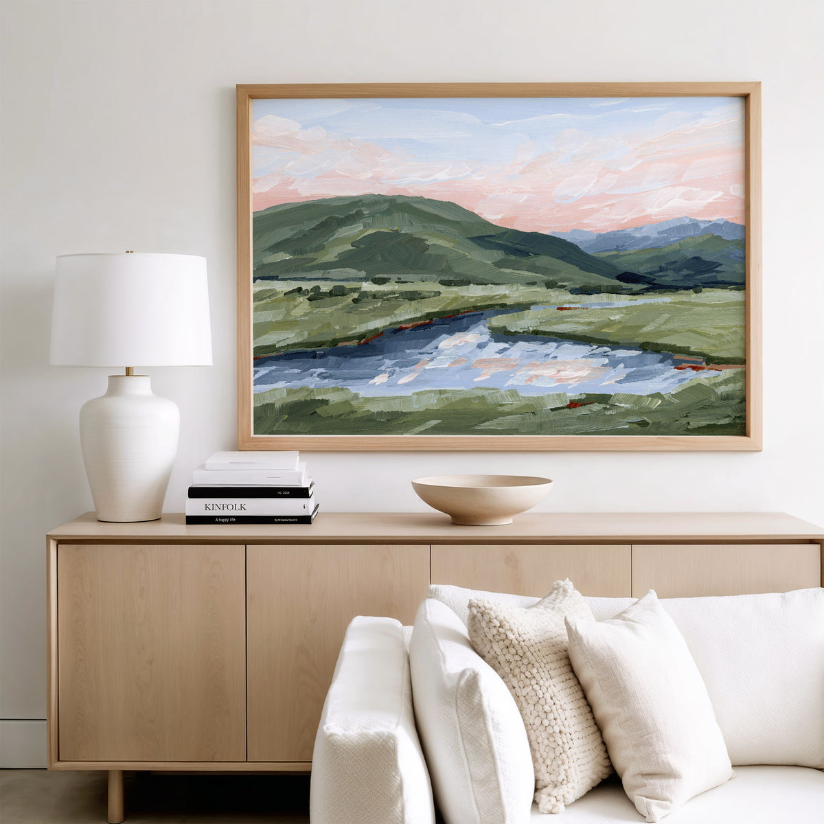 A framed landscape painting of green hills, a river, and a pink sunset sky, displayed on a light wood console above a white sofa in a modern living room setting.