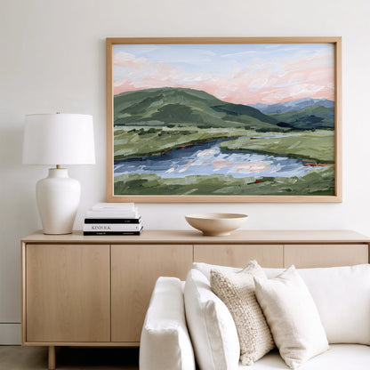 A framed landscape painting of green hills, a river, and a pink sunset sky, displayed on a light wood console above a white sofa in a modern living room setting.