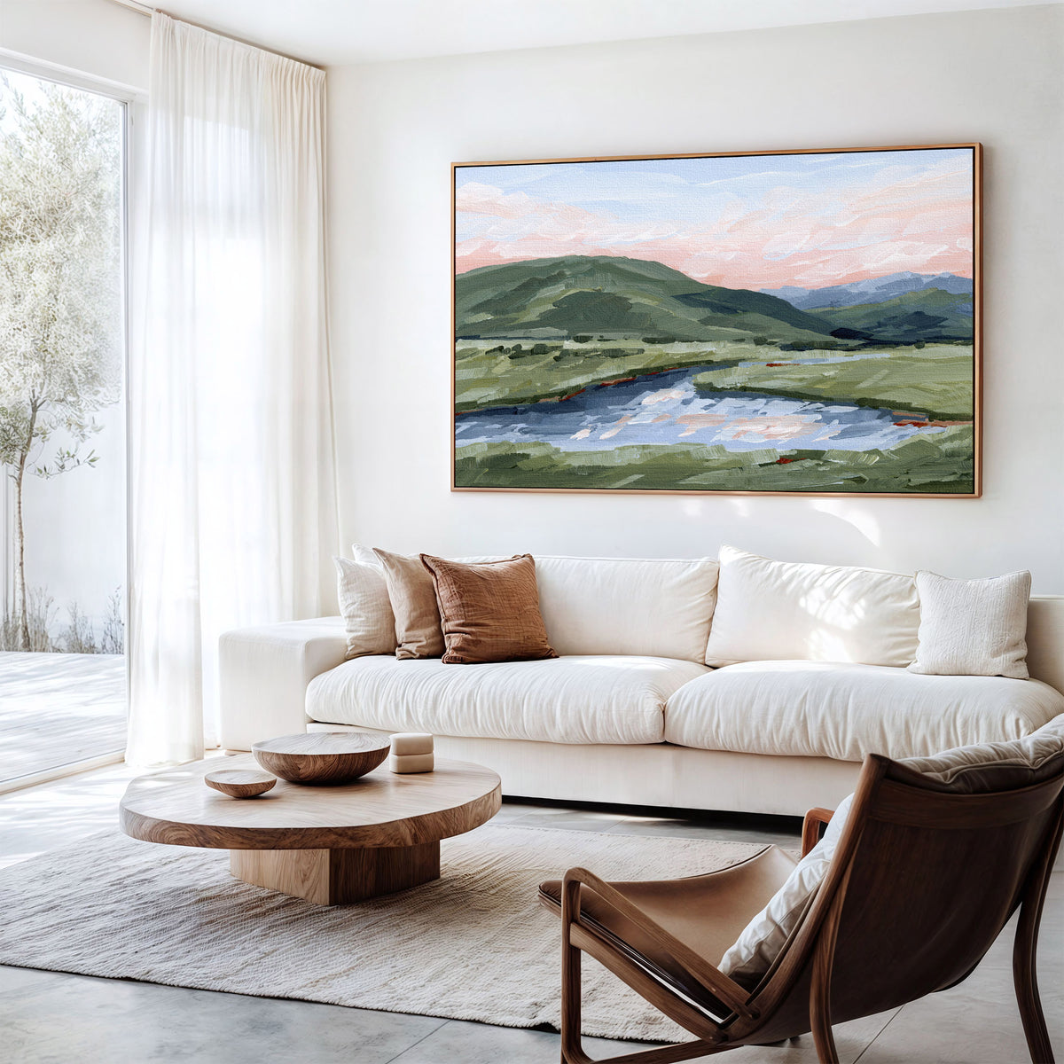 A large framed landscape painting of green hills and a reflective lake under a soft pink sky, displayed on a white wall above a cream sofa in a bright modern living room.