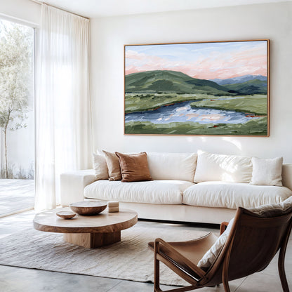 A large framed landscape painting of green hills and a reflective lake under a soft pink sky, displayed on a white wall above a cream sofa in a bright modern living room.