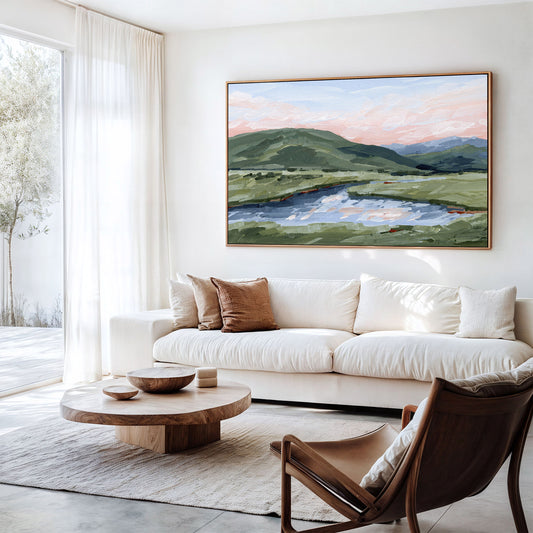 A large framed landscape painting of green hills and a reflective lake under a soft pink sky, displayed on a white wall above a cream sofa in a bright modern living room.