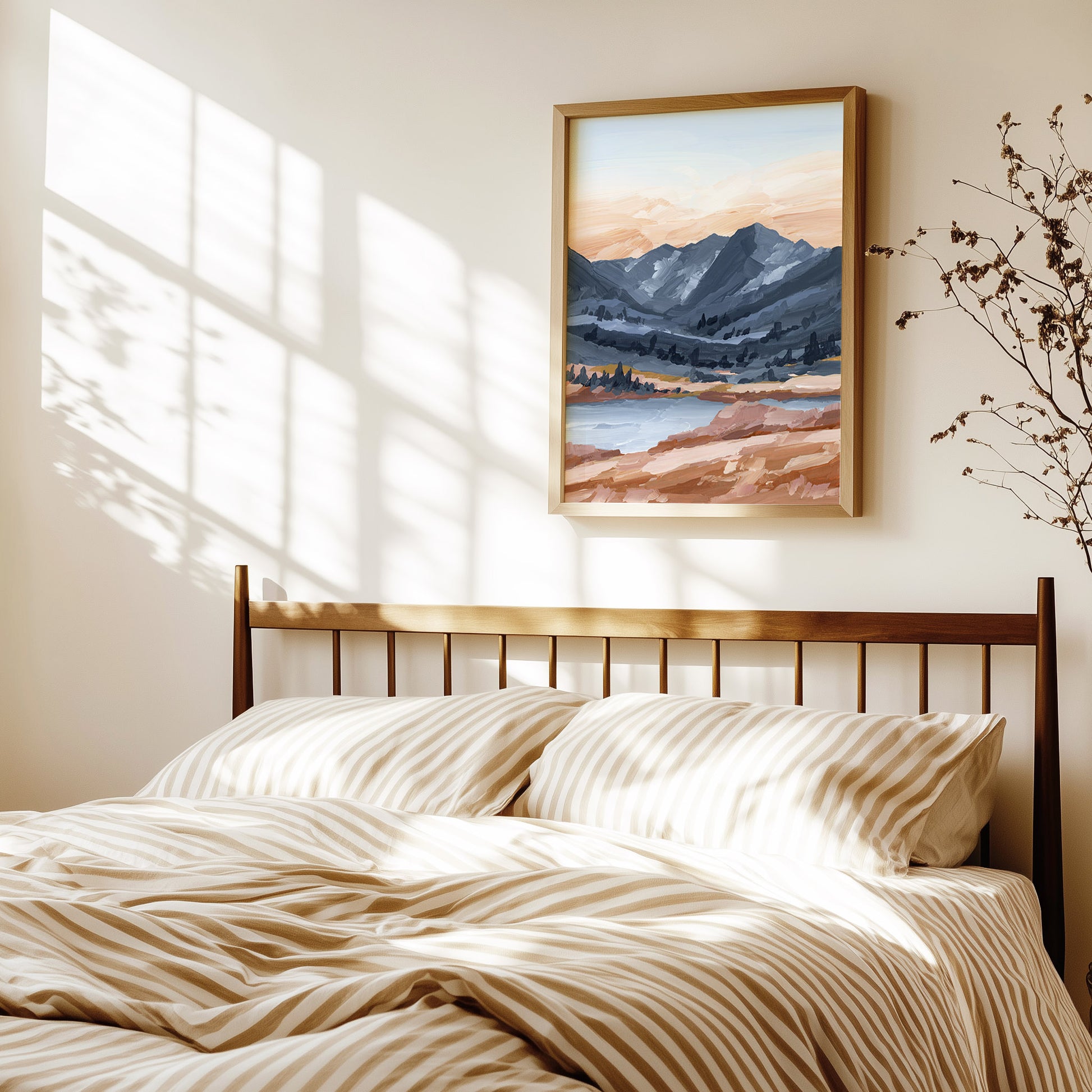 A framed landscape painting of mountains and a lake hangs on a beige wall above a bed with beige and white striped bedding, styled in a warm, sunlit bedroom.