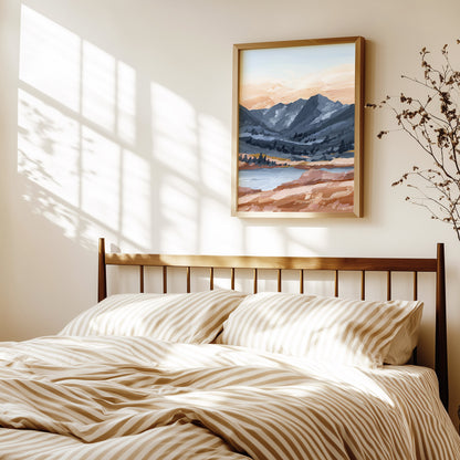 A framed landscape painting of mountains and a lake hangs on a beige wall above a bed with beige and white striped bedding, styled in a warm, sunlit bedroom.