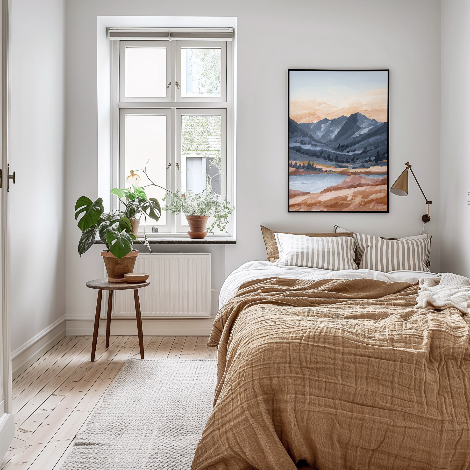 A framed landscape art print of mountains and a lake hangs on a light gray wall above a neatly made bed in a bright, modern bedroom with beige bedding, striped pillows, a window, and potted plants.