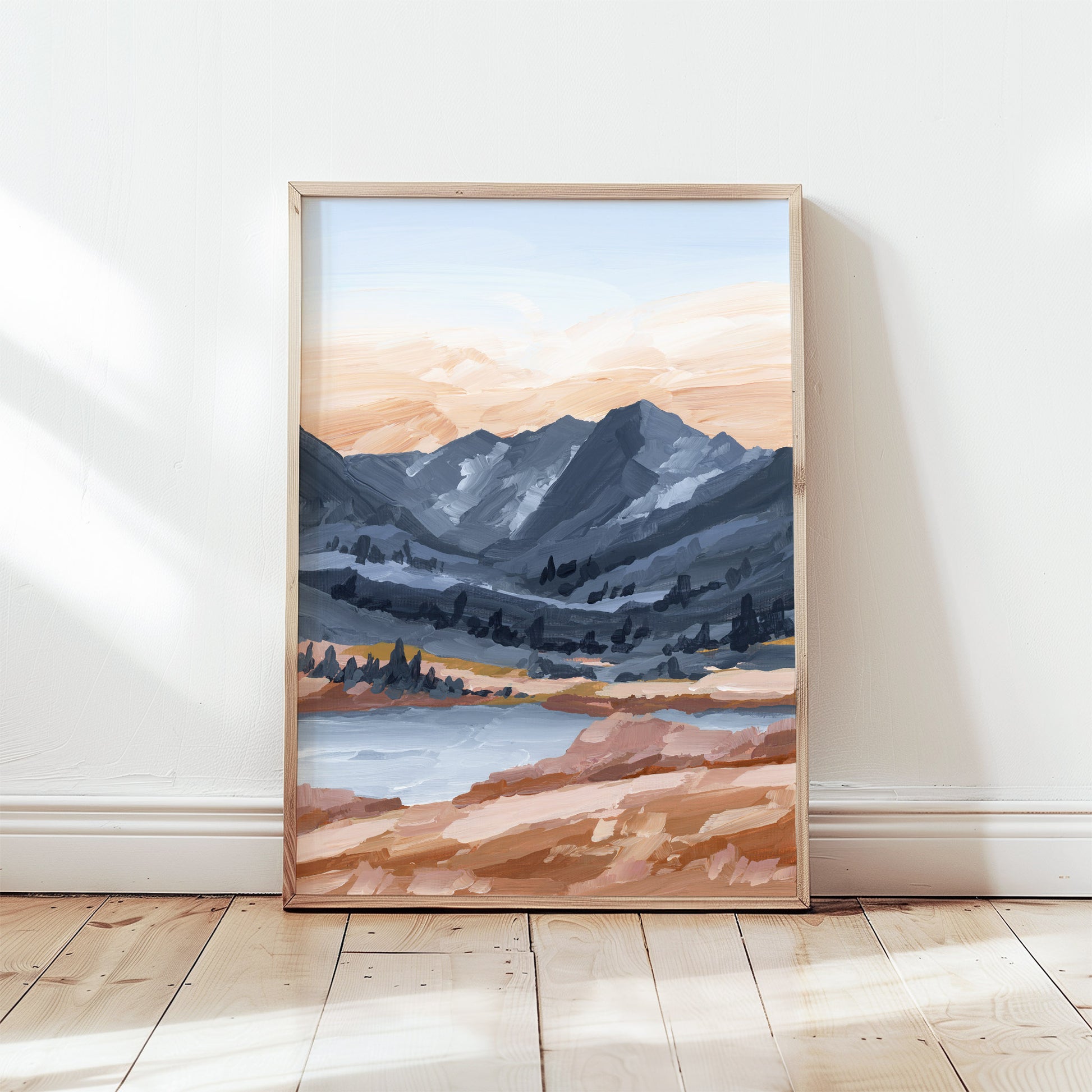 A framed landscape art print leaning against a white wall on a light wood floor. The painting depicts blue-gray mountains, a pale sky, and warm peach and brown foreground tones.