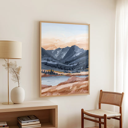 A framed landscape art print featuring blue-gray mountains, a lake, and warm peach and brown foreground tones, displayed on a cream wall in a styled room with a lamp, vase, and chair.