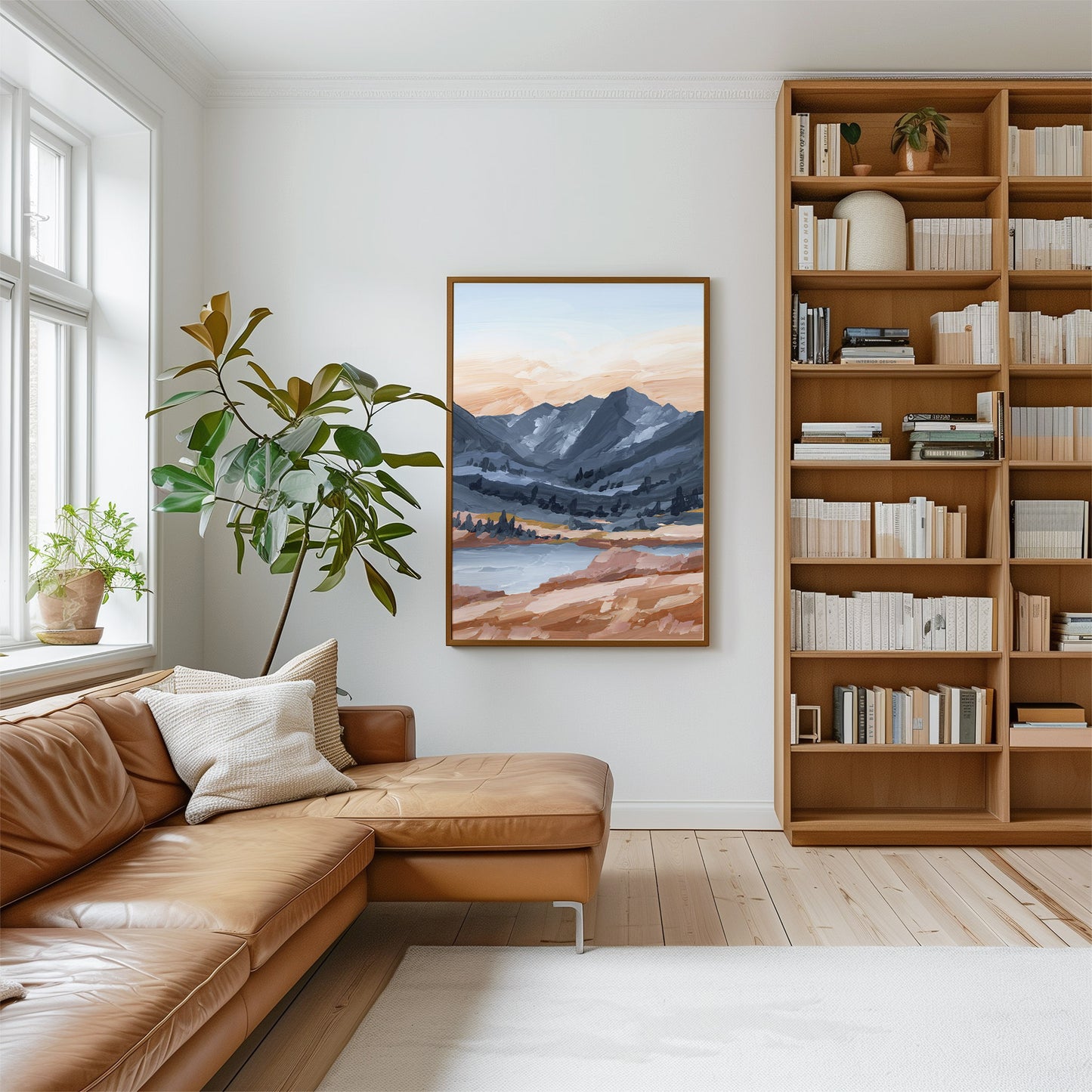 A framed landscape painting of a mountain valley at sunset hangs on a white wall in a bright living room with a brown leather sofa, a tall bookshelf, and a large potted plant.