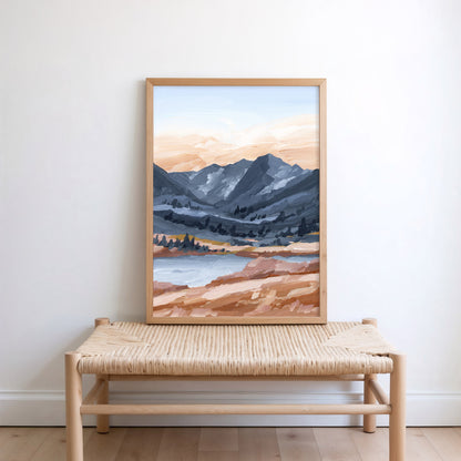 A framed landscape art print depicting mountains, a lake, and a sandy shoreline, leaning against a white wall on a light wood bench with a woven seat.