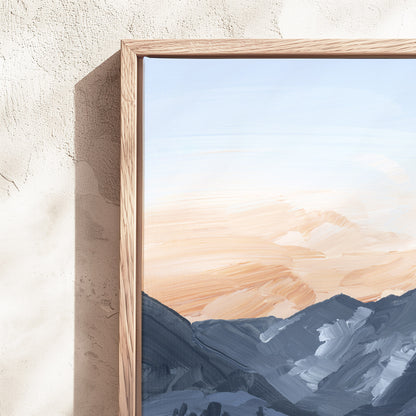 A framed landscape art print featuring textured brushstrokes of blue-gray mountains under a soft peach and cream sky, set in a light wood frame against a textured beige plaster wall.